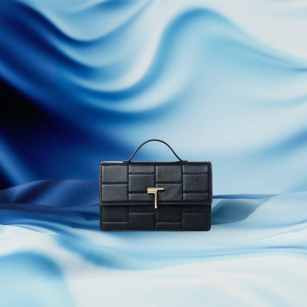 A black Trevony medium Minerva handbag in lambskin with a gold 'T' clasp, set against a flowing blue satin background.