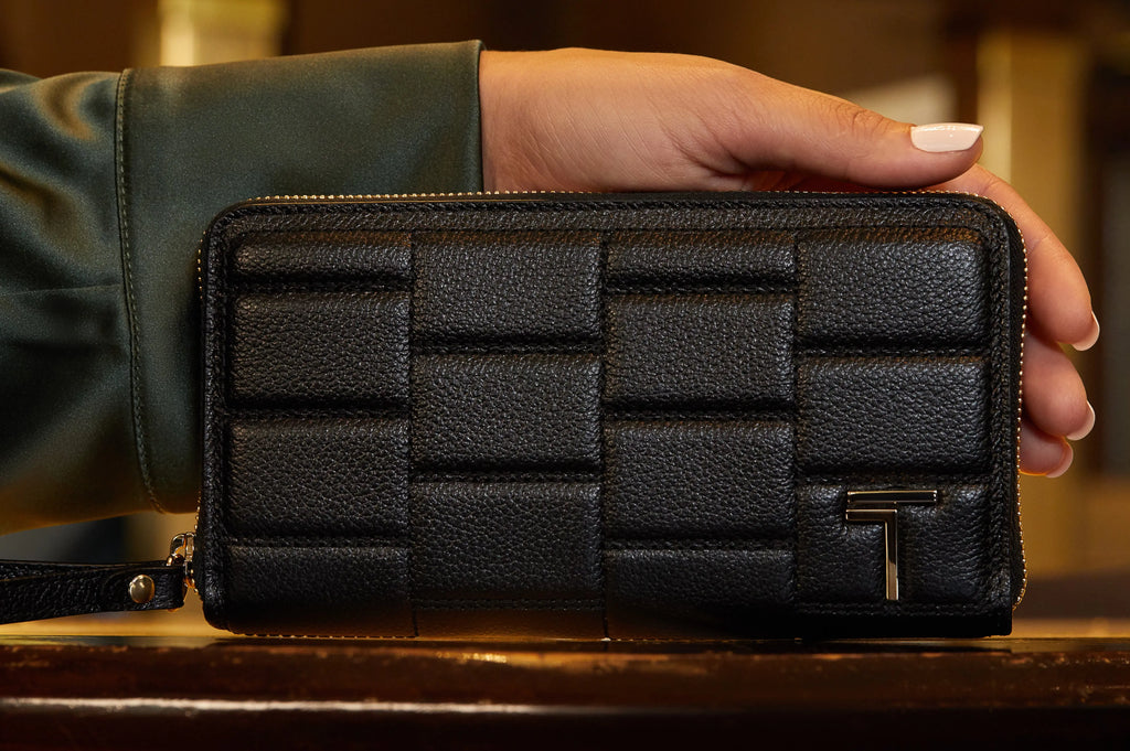 A hand holding a textured black Trevony Zipped wallet in lambskin with a prominent zipper and a metallic 'T' logo.