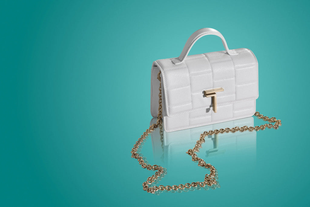 A white Trevony Minerva handbag with a lambskin exterior and gold hardware, including a chain crossbody strap, displayed against a turquoise background with a reflective surface.