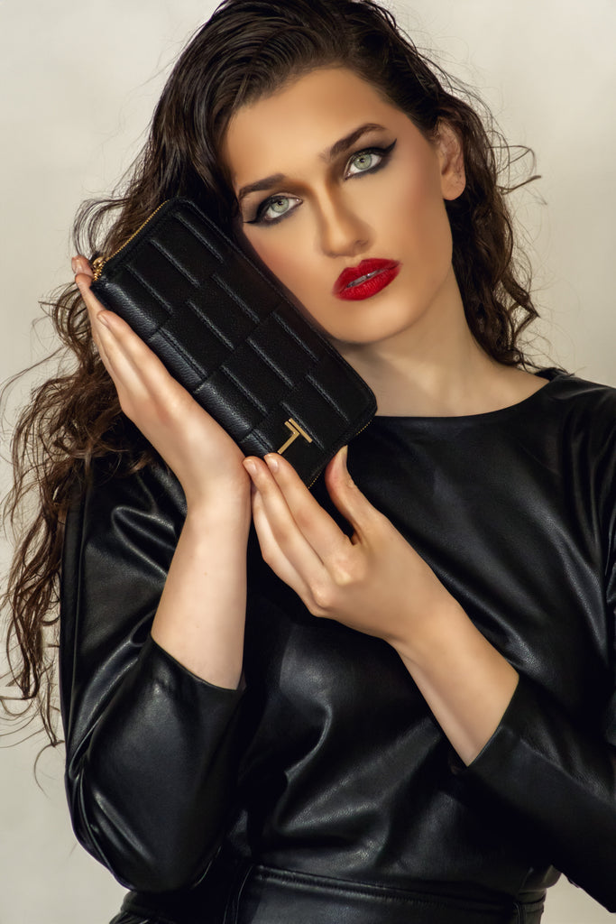 A close-up image of a model holding a black lambskin Trevony Zipped Wallet. The wallet has a textured pattern, and there is a golden T-shaped logo on the front.