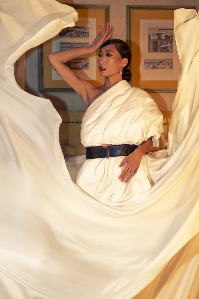 A person poses dramatically in a cream-colored, draped outfit accented with a Trevony Truth Oversized belt at the waist.
