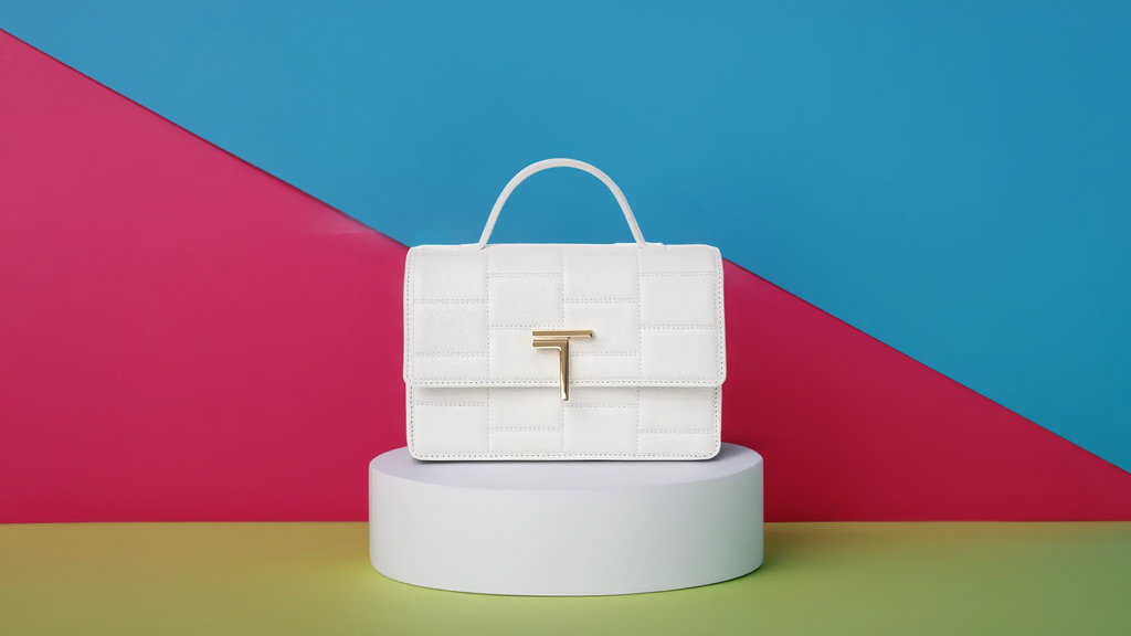 White textured Minerva handbag with a gold Trevony logo clasp, centered on a cylindrical pedestal with a bold geometric background in blue, pink, and yellow.