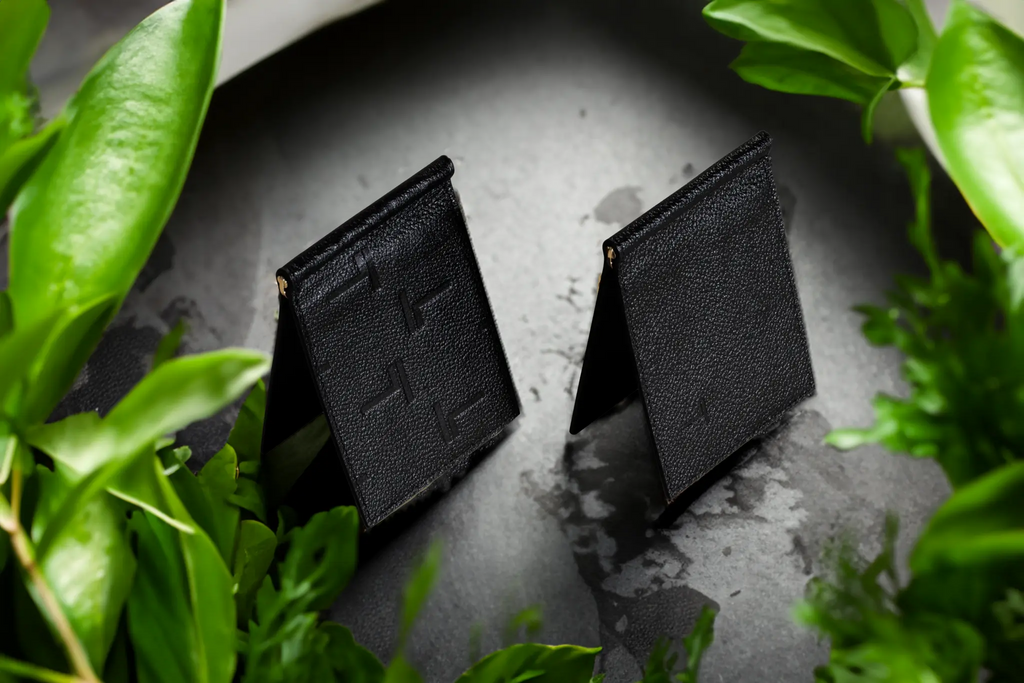 Two black textured lambskin leather Trevony wallets standing amidst lush green leaves on a speckled gray surface, showcasing the brand's embossed logo.