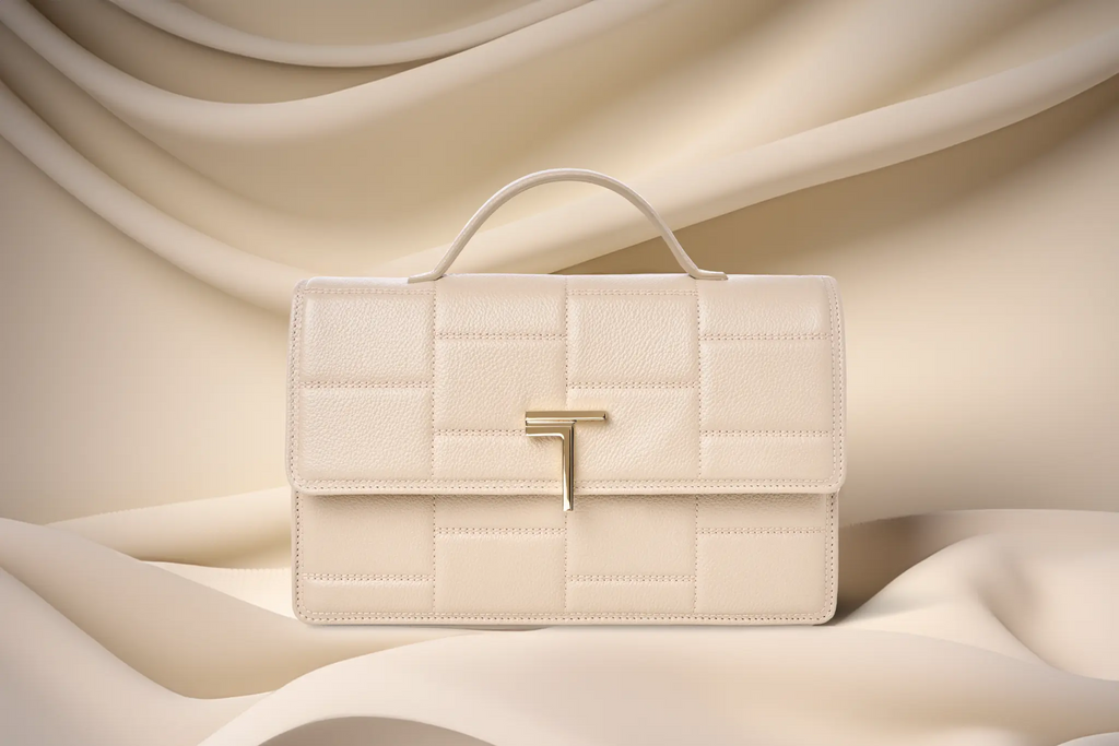 Elegant beige Trevony Minerva handbag with a textured finish, gold T logo closure, showcased against a flowing satin fabric backdrop in a complementary soft beige tone.